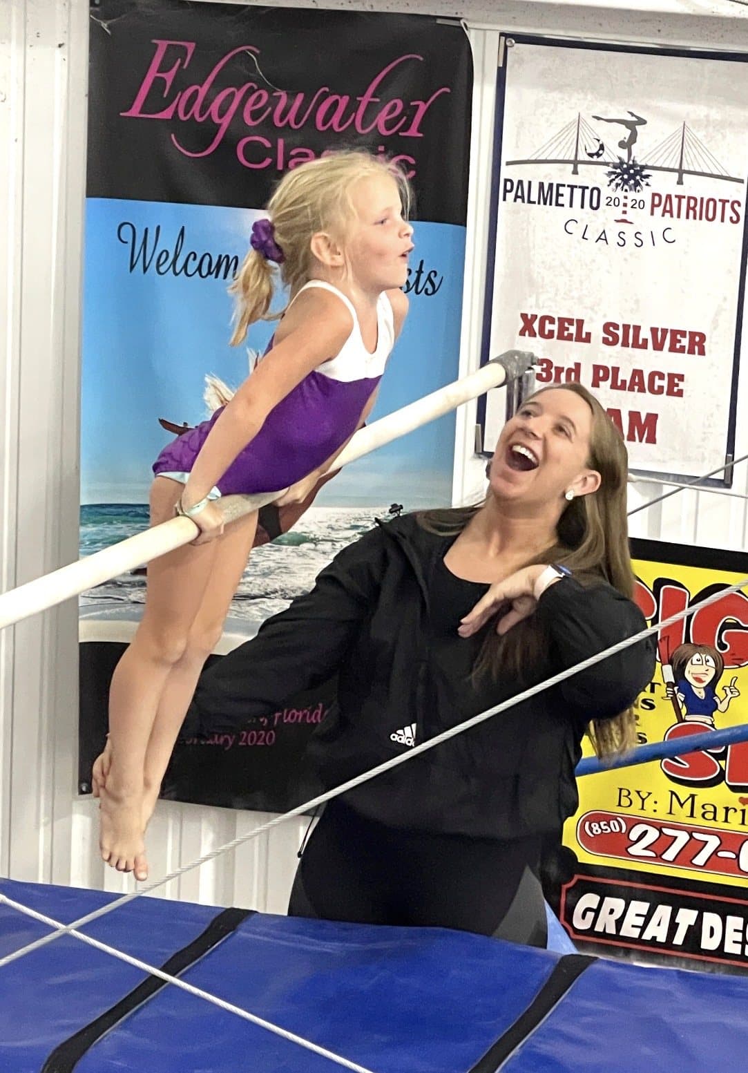 A smiling gymnast on the bar with her coach cheering her on